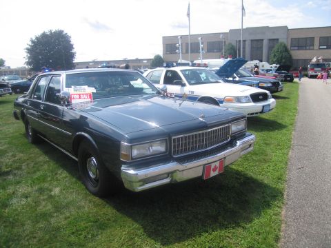 1991 Ford LTD Crown Victoria - dark gray Chevrolet Caprice four-door sedan with a "For Sale" sign in the window and a Canada flag plate, on grass.