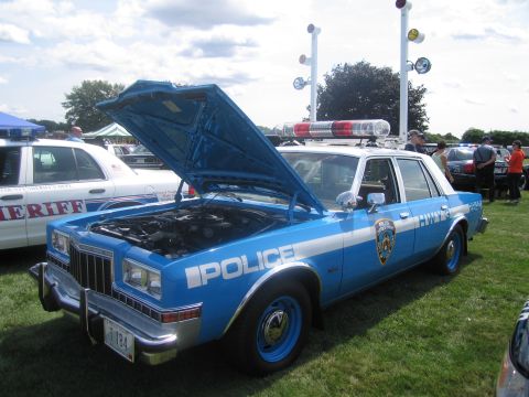 1991 Ford LTD Crown Victoria - blue full-size police sedan lettered "Police" with an NYPD-style shield, hood open and roof light bar, on grass.