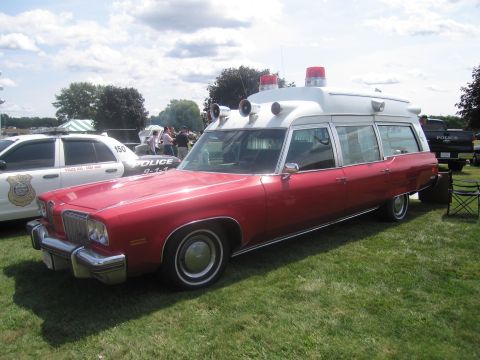 1991 Ford LTD Crown Victoria - vintage red-and-white Cadillac professional ambulance with roof-mounted lights and siren, displayed on grass at a show.