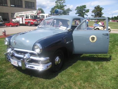 1991 Ford LTD Crown Victoria - Restored vintage two-tone blue Ford coupe styled as a 1951 state police car, door open with an agency seal, parked on grass by a fire truck.