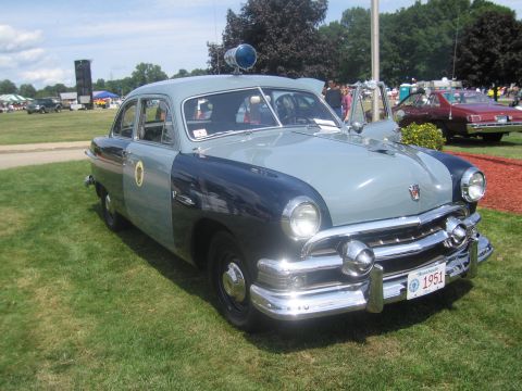 1991 Ford LTD Crown Victoria - Front three-quarter view of a restored two-tone blue vintage Ford police sedan with a roof spotlight and a 1951 plate, parked on grass.