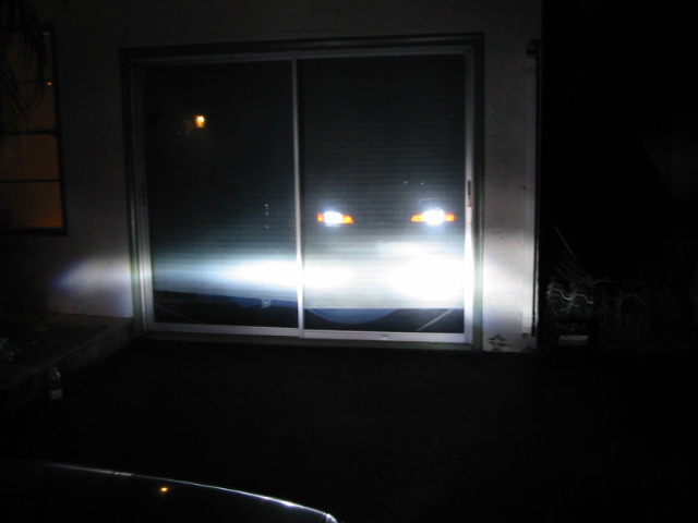 Car headlights shining brightly at a sliding glass door at night. Supermotors.net logo visible