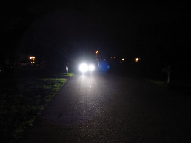 Car headlights illuminate a dark road at night, with a blue lens flare visible
