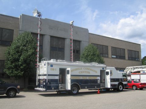 1991 Ford LTD Crown Victoria - white and blue Mobile Command Center truck with raised camera and antenna masts parked beside a fire apparatus outside a building.