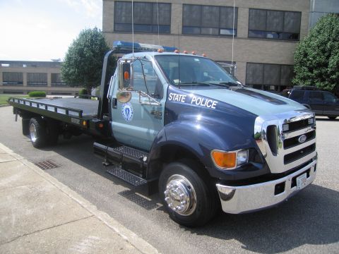 1991 Ford LTD Crown Victoria - Passenger-front three-quarter view of a two-tone blue Ford State Police flatbed tow truck parked outside a building.