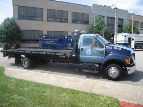 1991 Ford LTD Crown Victoria - Side view of a blue Ford State Police flatbed tow truck carrying a blue tactical operations equipment box on its bed.