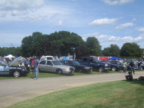 1991 Ford LTD Crown Victoria - lineup of police vehicles including a state police sedan, Crown Victoria, and SUV with hoods up, displayed on grass at an outdoor show.