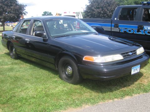 1991 Ford LTD Crown Victoria - black Ford Crown Victoria police sedan with steel wheels parked on grass next to a blue UMass Police pickup truck.