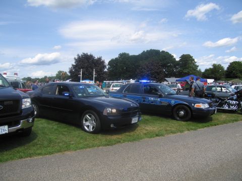 1991 Ford LTD Crown Victoria - dark Dodge Charger police sedan beside a blue UMass Police Crown Victoria with light bars, lined up on grass under a cloudy sky.