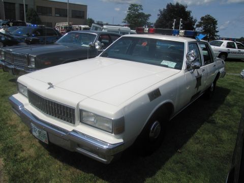 1991 Ford LTD Crown Victoria - Front-three-quarter view of a white boxy Chevrolet Caprice police sedan with spotlight and light bar, parked on grass at a show.