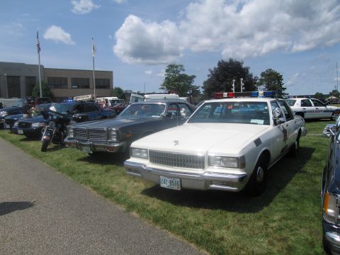 1991 Ford LTD Crown Victoria - lineup of classic police vehicles on grass led by a white Chevrolet Caprice cruiser, with older sedans and a motorcycle behind.