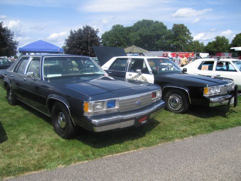 1991 Ford LTD Crown Victoria - dark Ford LTD Crown Victoria four-door sedan beside a black-and-white police version, parked on grass at an outdoor show.