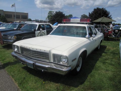 1991 Ford LTD Crown Victoria - white Plymouth Fury full-size four-door police sedan with roof light bar, parked on grass among other classic cruisers.