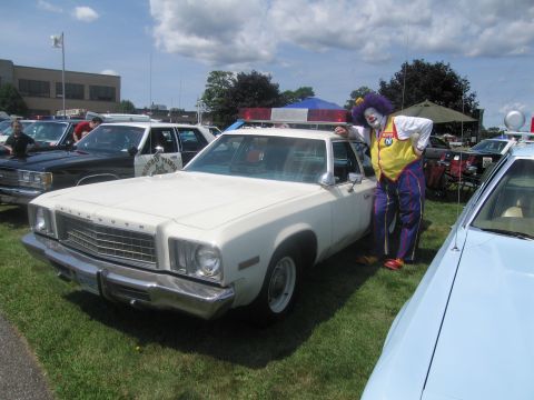 1991 Ford LTD Crown Victoria - person in a colorful clown costume stands beside a white Plymouth Fury full-size sedan on grass at an outdoor car show.