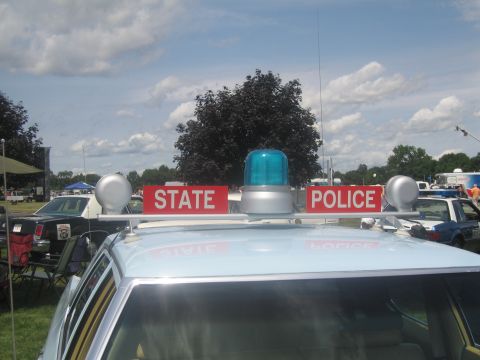 1991 Ford LTD Crown Victoria - Roof detail of a cruiser showing red STATE POLICE signs flanking a blue dome light and spotlights, against a cloudy sky.