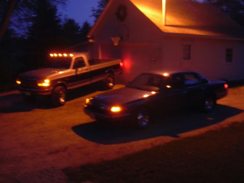 1996 Ford Crown Victoria - A dark sedan and a pickup truck are parked in front of a house at night, with their lights on.
