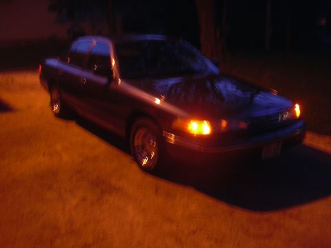 1996 Ford Crown Victoria - A dark sedan is parked on a paved surface at night with its headlights illuminated.