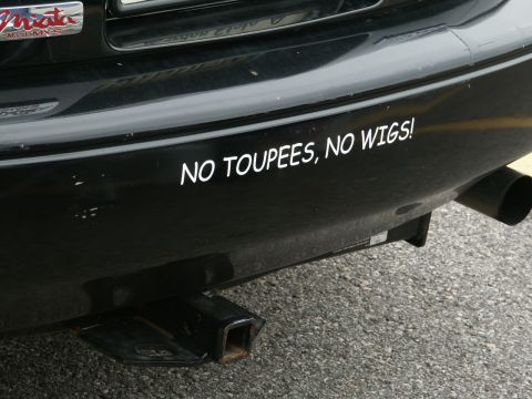 2001 Mercury Grand Marquis - A black car's rear bumper displays a humorous sticker and a trailer hitch, with a visible exhaust pipe.