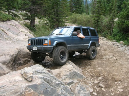 1995 Ford Bronco - A blue Jeep Cherokee XJ tackles a challenging rocky trail in a wooded mountain area.