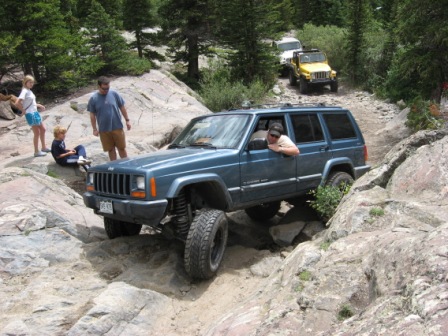1995 Ford Bronco - A blue Jeep Cherokee XJ navigates a challenging rocky off-road trail, with other vehicles and onlookers in the background.