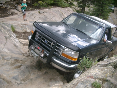 1995 Ford Bronco - A black 1995 Ford Bronco is shown off-roading on a rocky trail, with another vehicle visible in the distance.