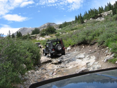 1995 Ford Bronco - Multiple Jeep Wranglers traverse a challenging, rocky mountain path under a partly cloudy sky.
