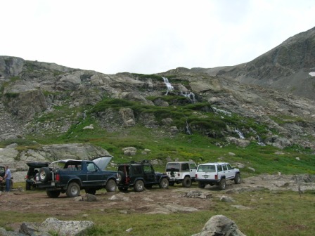 1995 Ford Bronco - Several off-road vehicles are parked on a mountain trail with waterfalls cascading down the rocky slope.