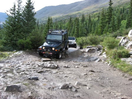 1995 Ford Bronco - Two SUVs tackle a challenging, rocky mountain trail surrounded by dense pine forest.