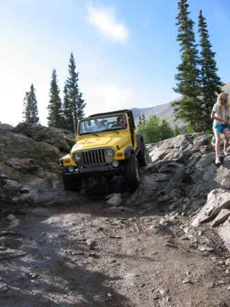 1995 Ford Bronco - A yellow Jeep navigates a challenging rocky terrain on a sunny day with trees and mountains in the background.