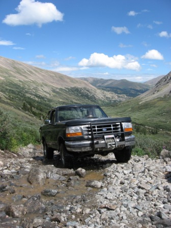 1995 Ford Bronco - A black Ford Bronco navigates a rocky mountain path under a blue sky with scattered clouds.