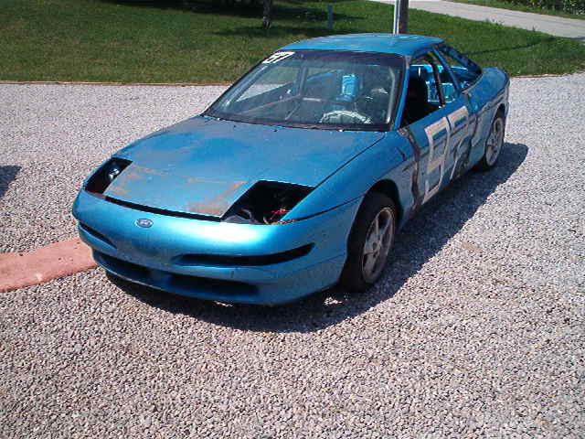 Blue race car with numbers on the side, parked on gravel. Hood is open and headlights are missing