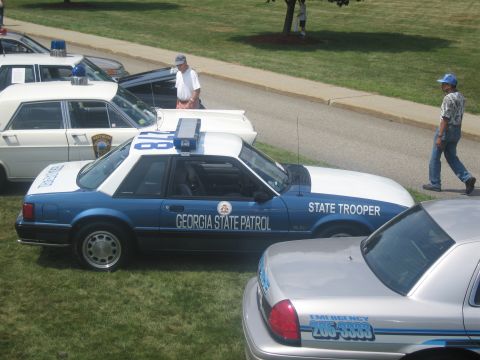 1991 Ford LTD Crown Victoria - blue and white Ford Mustang lettered GEORGIA STATE PATROL and STATE TROOPER sits on grass beside other cruisers at a show.