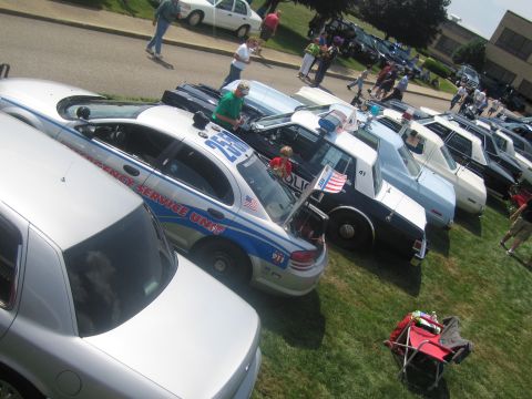 1991 Ford LTD Crown Victoria - From above, several police cruisers including an Emergency Service Unit car are lined up on grass with small flags and onlookers.