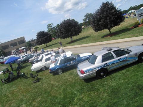 1991 Ford LTD Crown Victoria - high-angle shot of police cruisers parked diagonally on grass, including a silver POLICE car, with shade trees and a building in back.