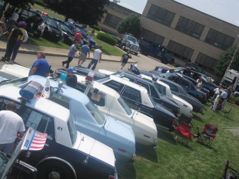 1991 Ford LTD Crown Victoria - long row of classic police sedans, including a black-and-white cruiser, is displayed on grass as people stroll past a brick building.
