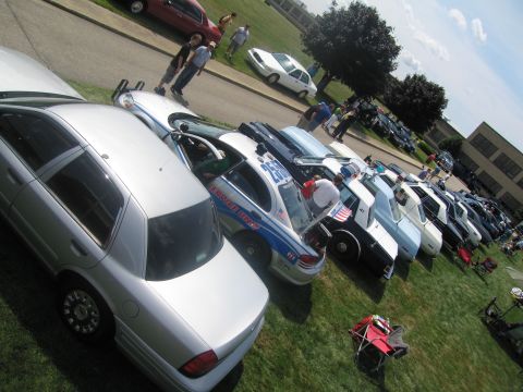 1991 Ford LTD Crown Victoria - angled view of police cars parked nose-out on grass, including a marked cruiser, with attendees walking on the pavement behind.
