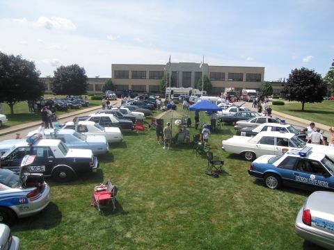 1991 Ford LTD Crown Victoria - wide overview of a police vehicle show, with cruisers parked on a lawn, tents and spectators in front of a long brick building.