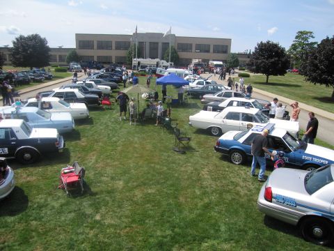 1991 Ford LTD Crown Victoria - high vantage shows numerous police cars arranged on a green lawn with canopy tents and visitors before a large brick building.