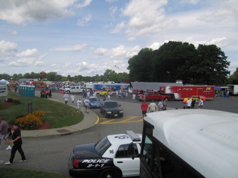 1991 Ford LTD Crown Victoria - busy outdoor event lot with race transporters and crowds, with a black-and-white POLICE 9-1-1 Crown Victoria parked in the foreground.