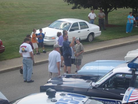 1991 Ford LTD Crown Victoria - group of adults and children gathers around police cruisers on a roadway, with a white Crown Victoria parked on the grass behind.