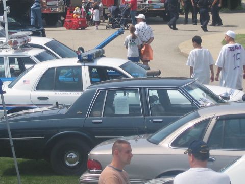 1991 Ford LTD Crown Victoria - Attendees walk among displayed police sedans, including a white Hampden County cruiser with a window placard, at an outdoor event.
