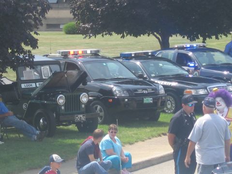 1991 Ford LTD Crown Victoria - black Hyundai Tucson SUV sits among police vehicles, including a SHERIFF unit and a vintage Jeep, as a clown and spectators look on.