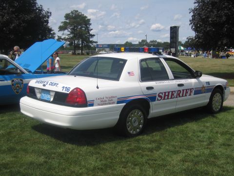 1991 Ford LTD Crown Victoria - white Ford Crown Victoria marked SHERIFF with red and blue striping sits on grass beside a blue NYPD cruiser at a car show.