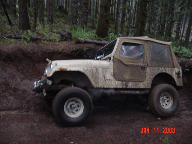 Tan Jeep CJ with soft top, large mud tires and lift, side view in muddy forest pit