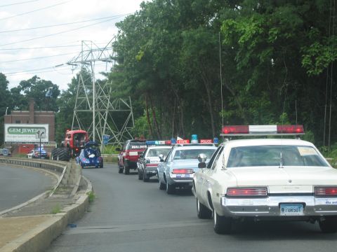 1991 Ford LTD Crown Victoria - procession of police cruisers and a red fire truck travels along a roadside past a Gem Jewelry building under power lines.