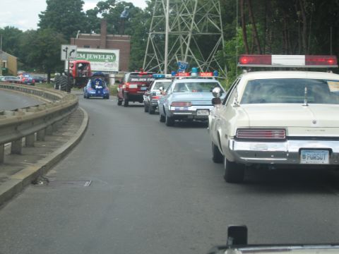 1991 Ford LTD Crown Victoria - cream-colored Buick with rooftop lights leads a parade of police cruisers along a road past a Gem Jewelry building.