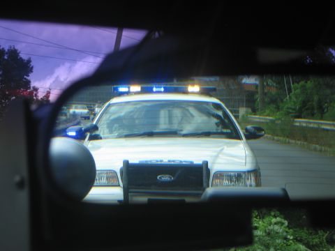 1991 Ford LTD Crown Victoria - white Ford police car with its blue lightbar lit approaches at dusk, framed through the rear window of another vehicle.