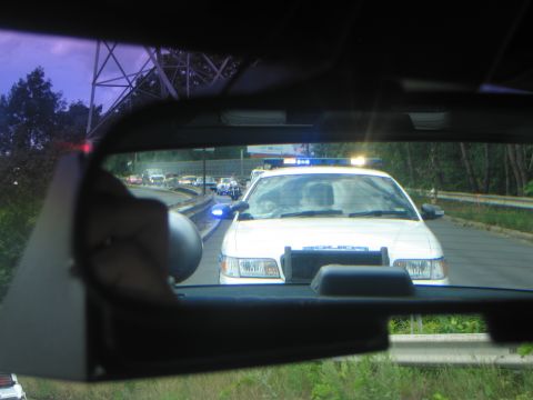 1991 Ford LTD Crown Victoria - white police cruiser with active lights follows behind, seen reflected in the rearview mirror of a moving vehicle.