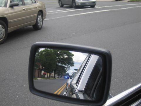 1991 Ford LTD Crown Victoria - car side mirror reflects a police vehicle with a blue light on a two-lane city street, with a tan sedan passing nearby.