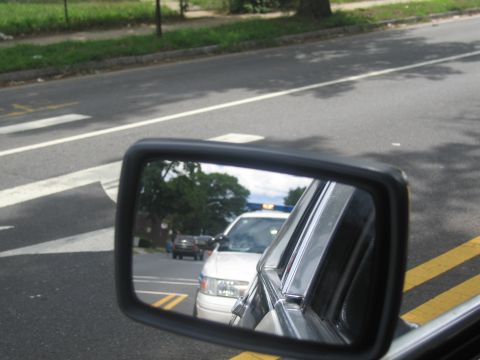 1991 Ford LTD Crown Victoria - vehicle side mirror reflects a white police car with a blue light following behind on a wide urban roadway.